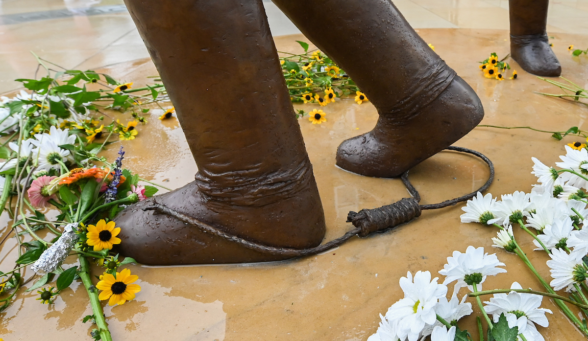 Bronze feet sculpture with a rope, surrounded by daisies and sunflowers on a wet surface.