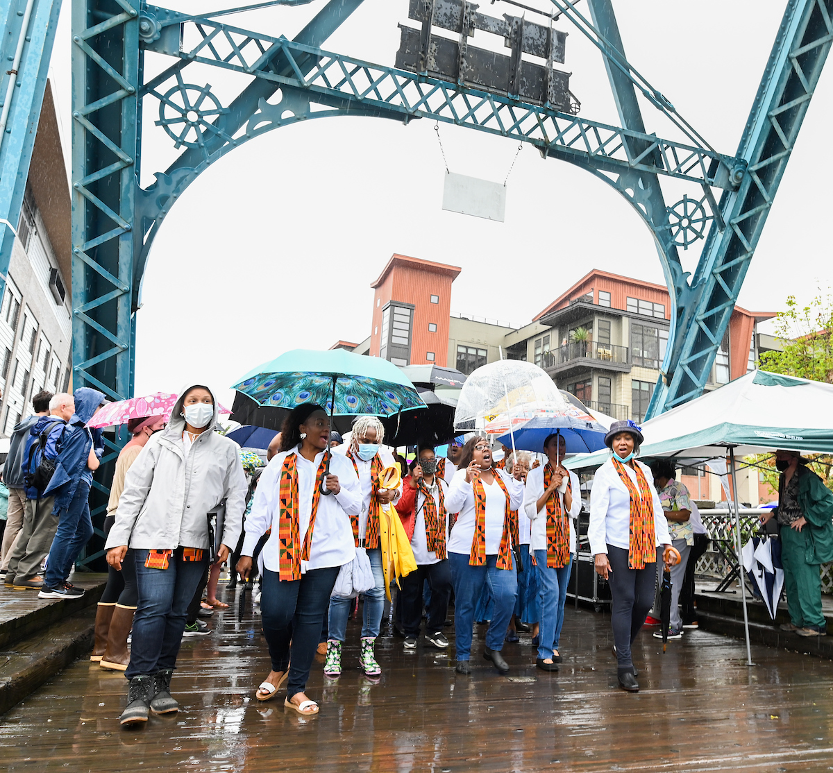 A group of people walks across a blue steel bridge in the rain, wearing colorful sashes and some holding umbrellas.