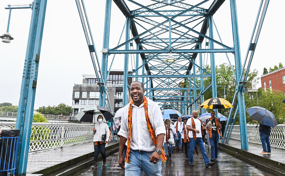 A group of people walks across a blue steel bridge in the rain, wearing colorful sashes and some holding umbrellas.