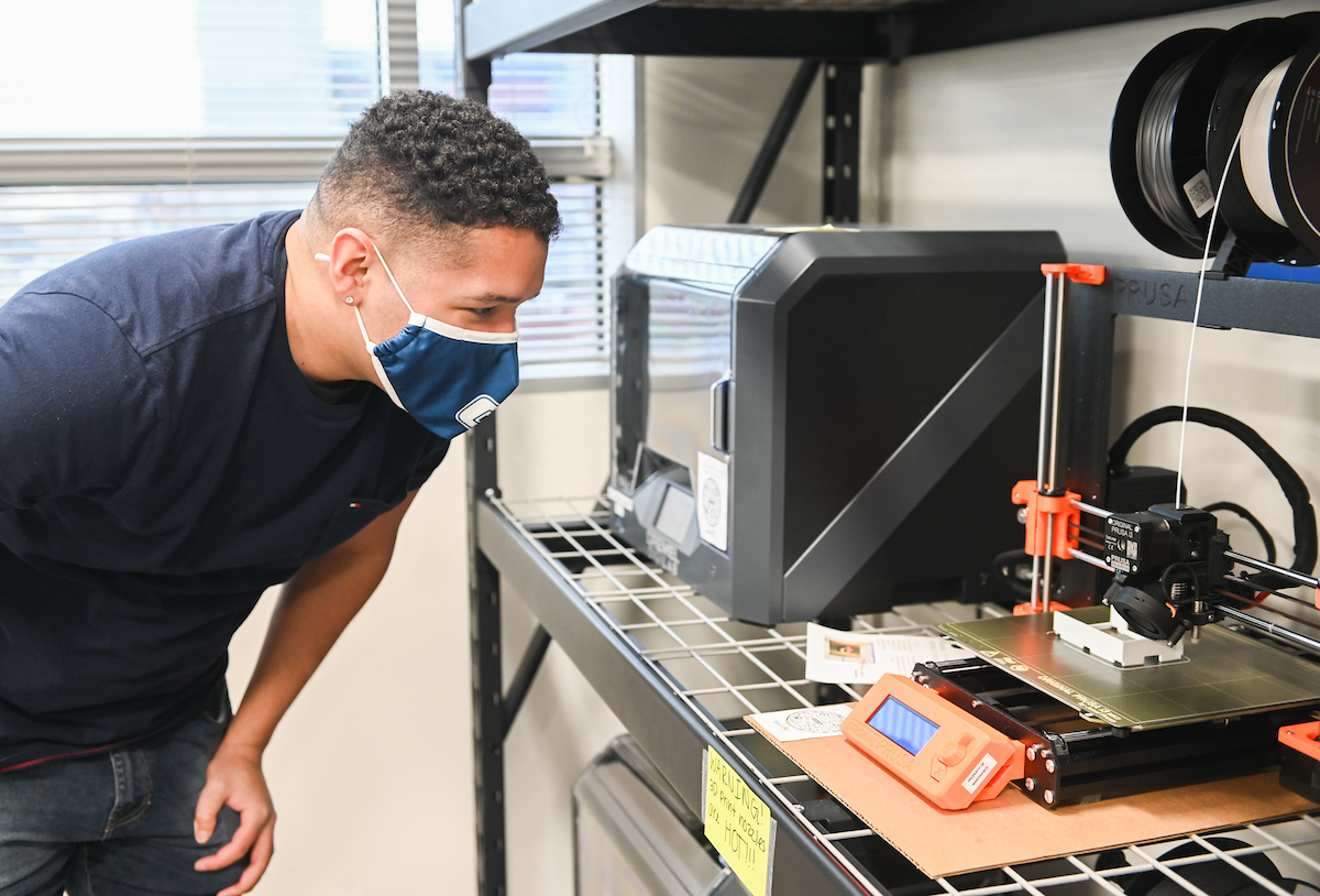 A student leans in to observe 3D printer in a workshop with metal shelves.