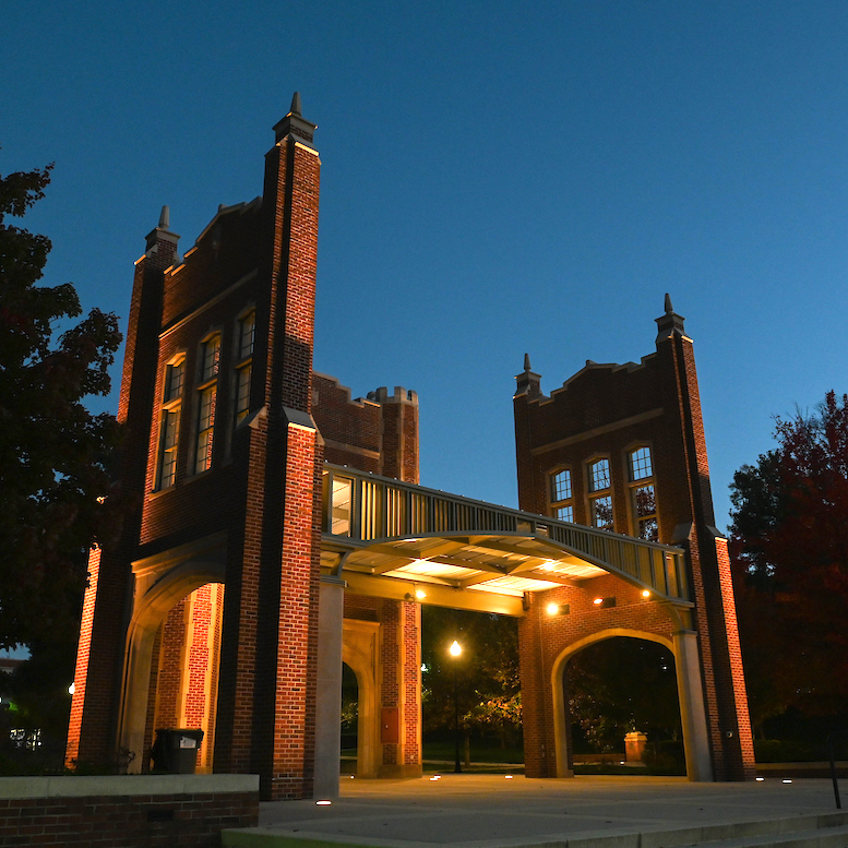 Illuminated brick archway with tall, pointed towers against a twilight sky.