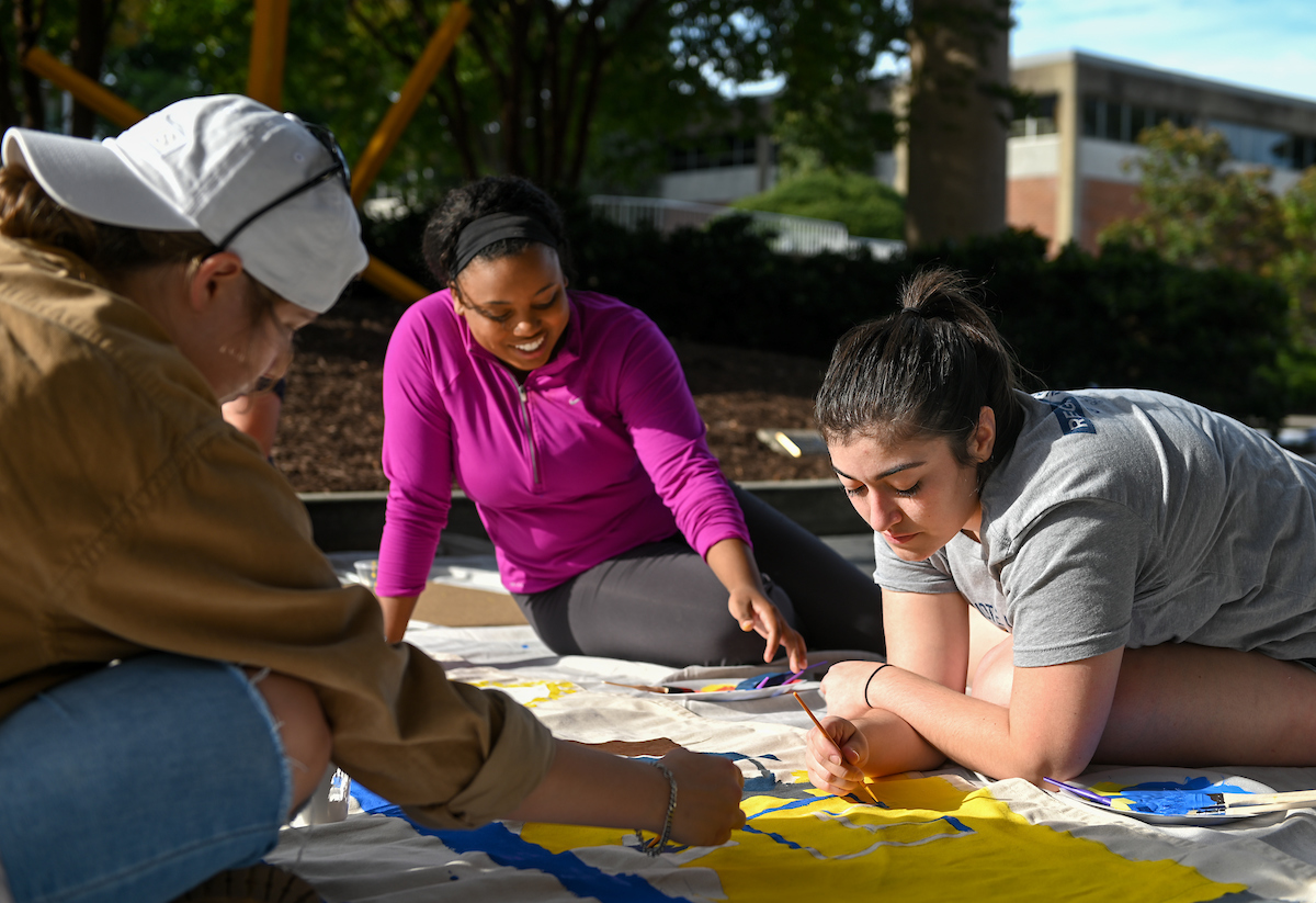 Three students are painting a large banner outdoors on a sunny day.