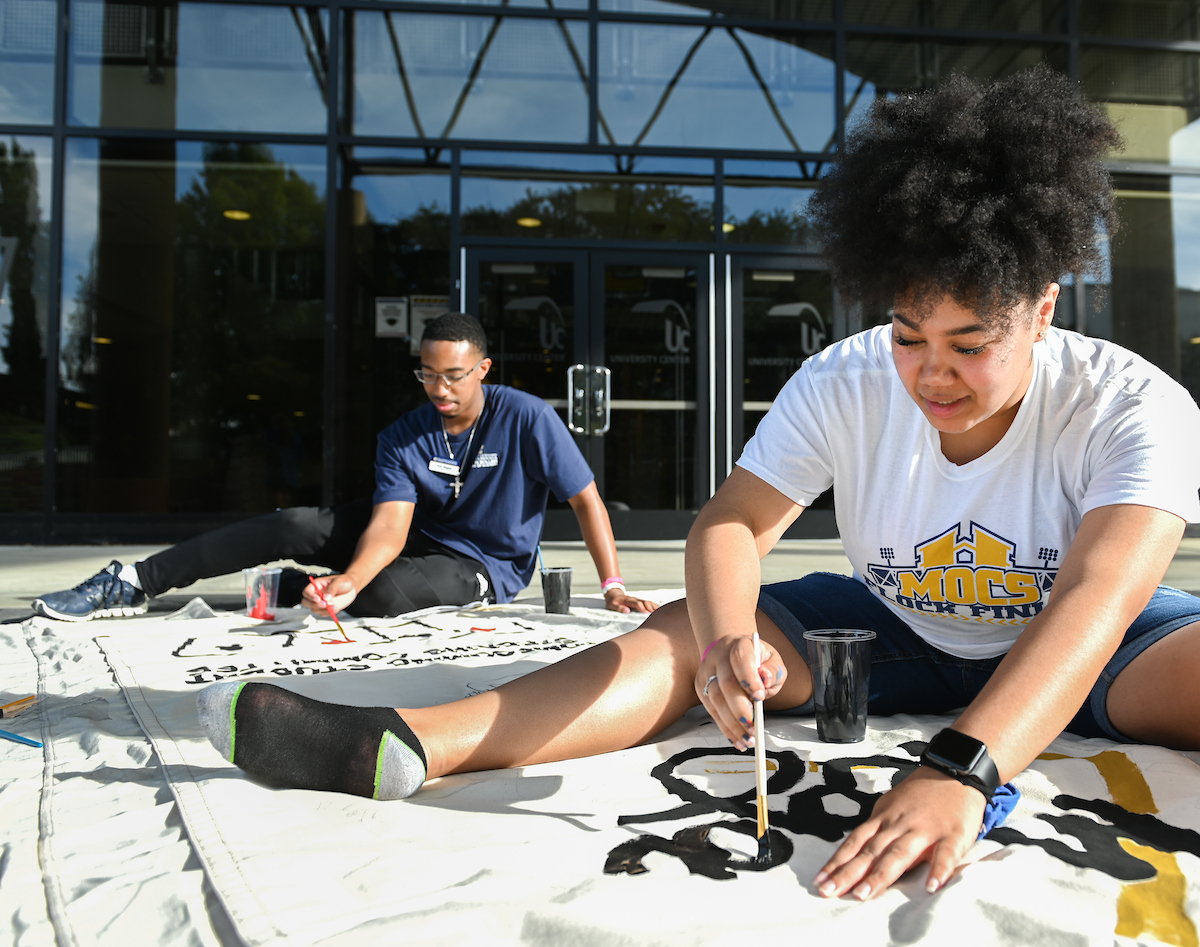 Two people sit outside painting a banner.