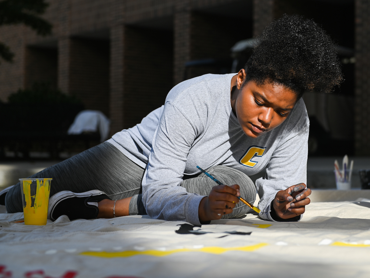 A student in a gray shirt concentrates intently, painting on a banner outdoors.