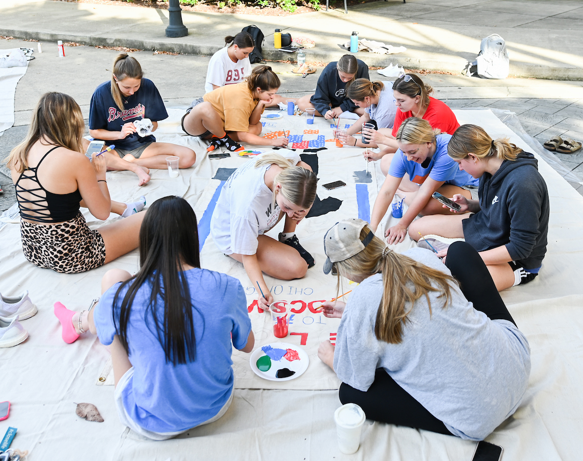 A group of students sitting on a large banner outdoors, painting.