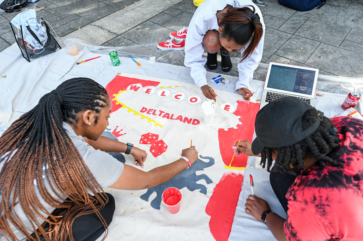 Three students paint a banner on the ground reading "Welcome Deltaland".