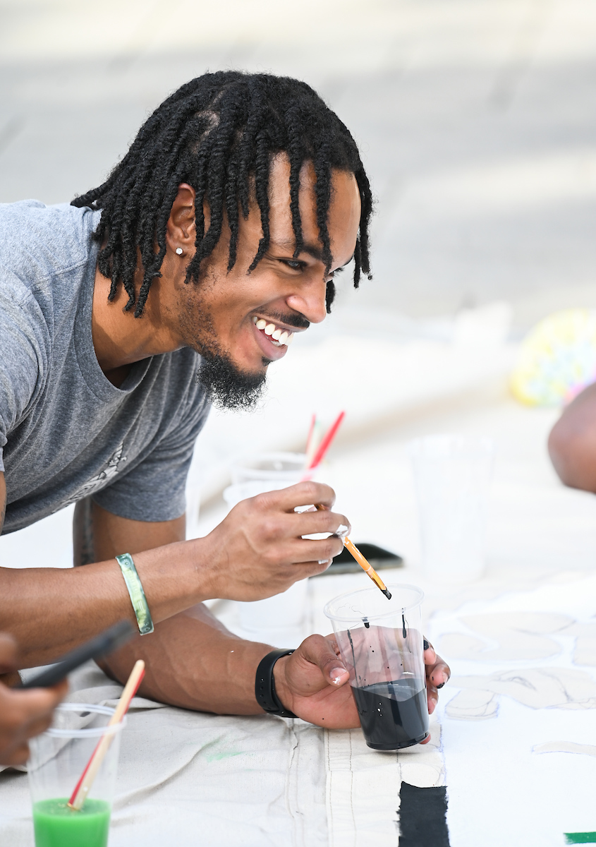 A student smiles while holding a paintbrush and a cup of paint.