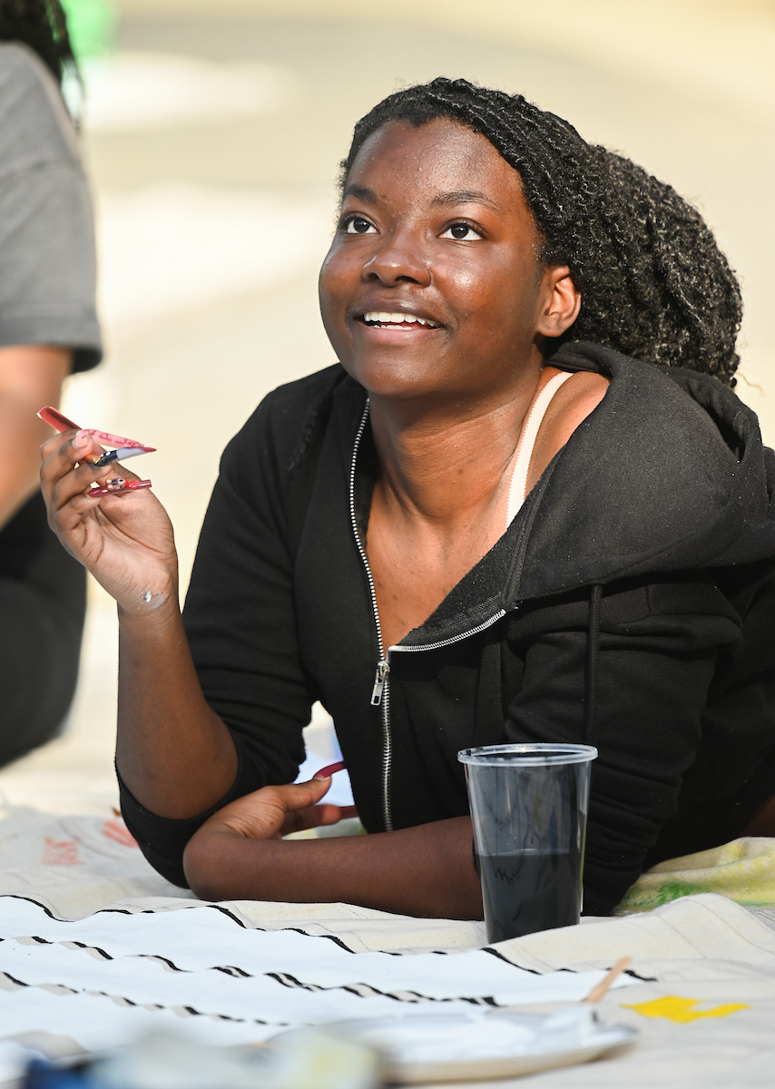A student smiles, holding a paintbrush and looking upward. A plastic cup with black liquid inside is nearby.