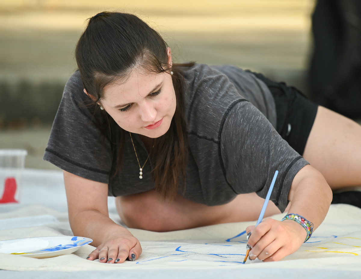 A student leans over while painting with a blue brush on a large banner on the floor.