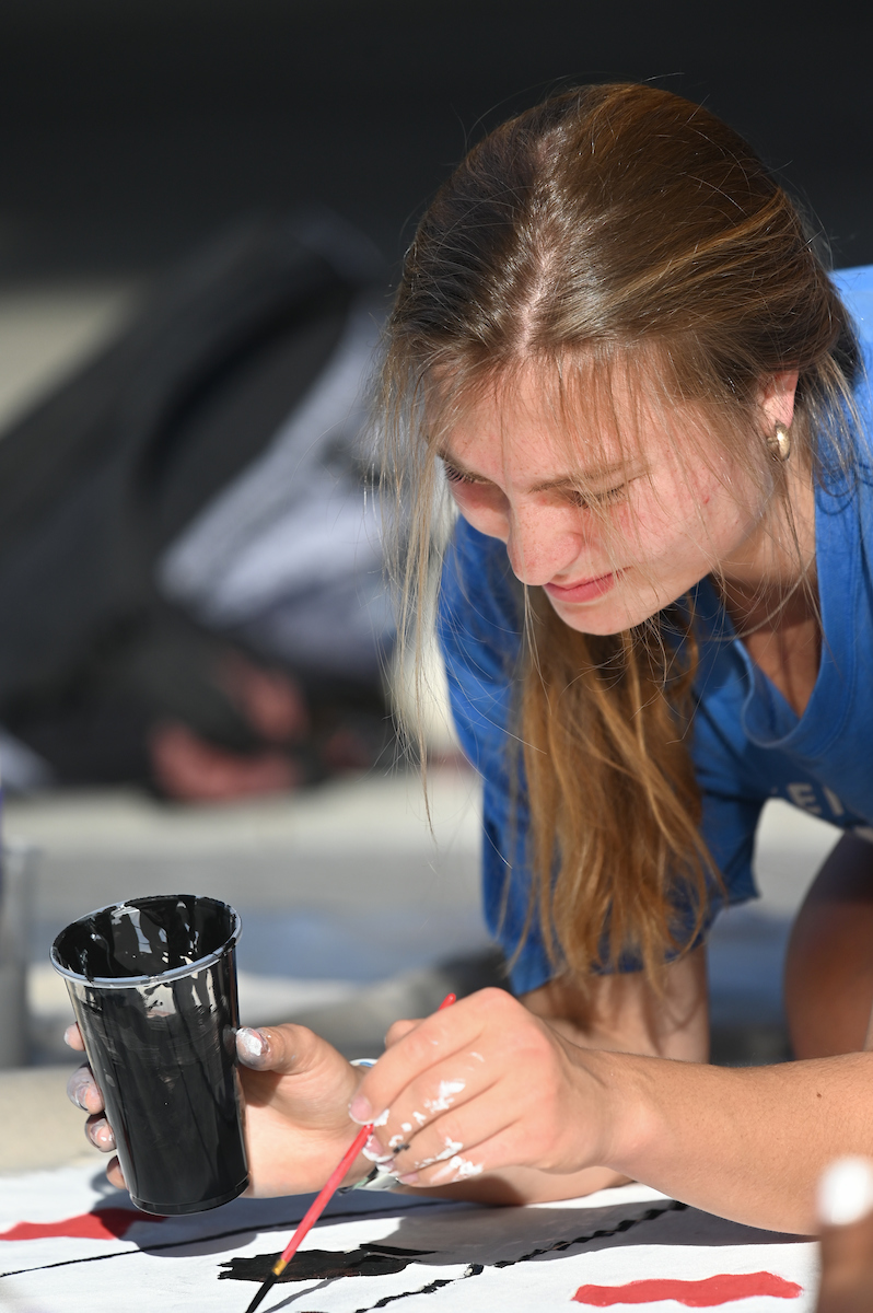 A student focuses while painting on a large banner. She holds a brush dipped in black paint, with a cup beside her.