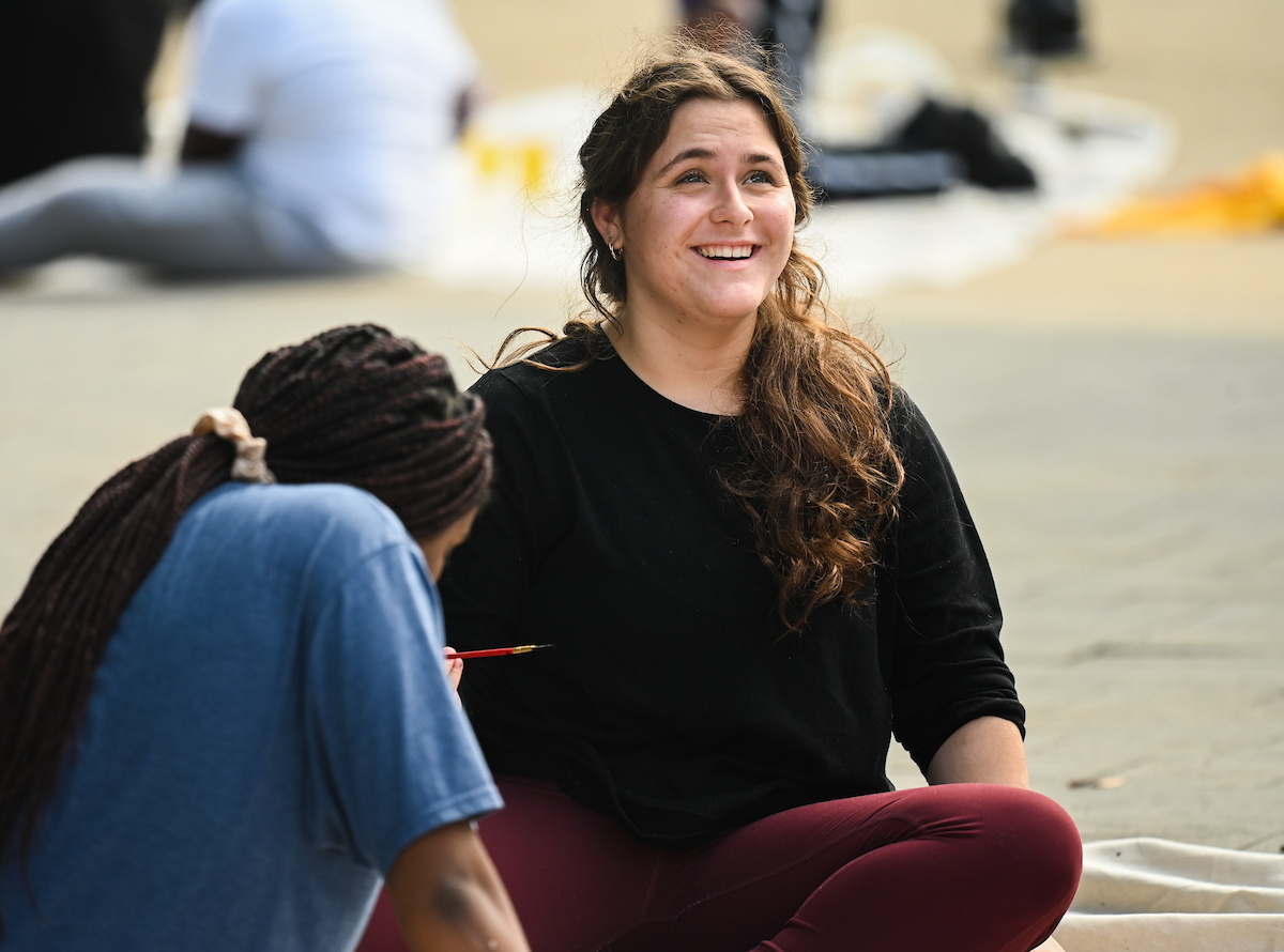 A woman sits on a street, smiling upward. Another person, partially visible in the foreground.