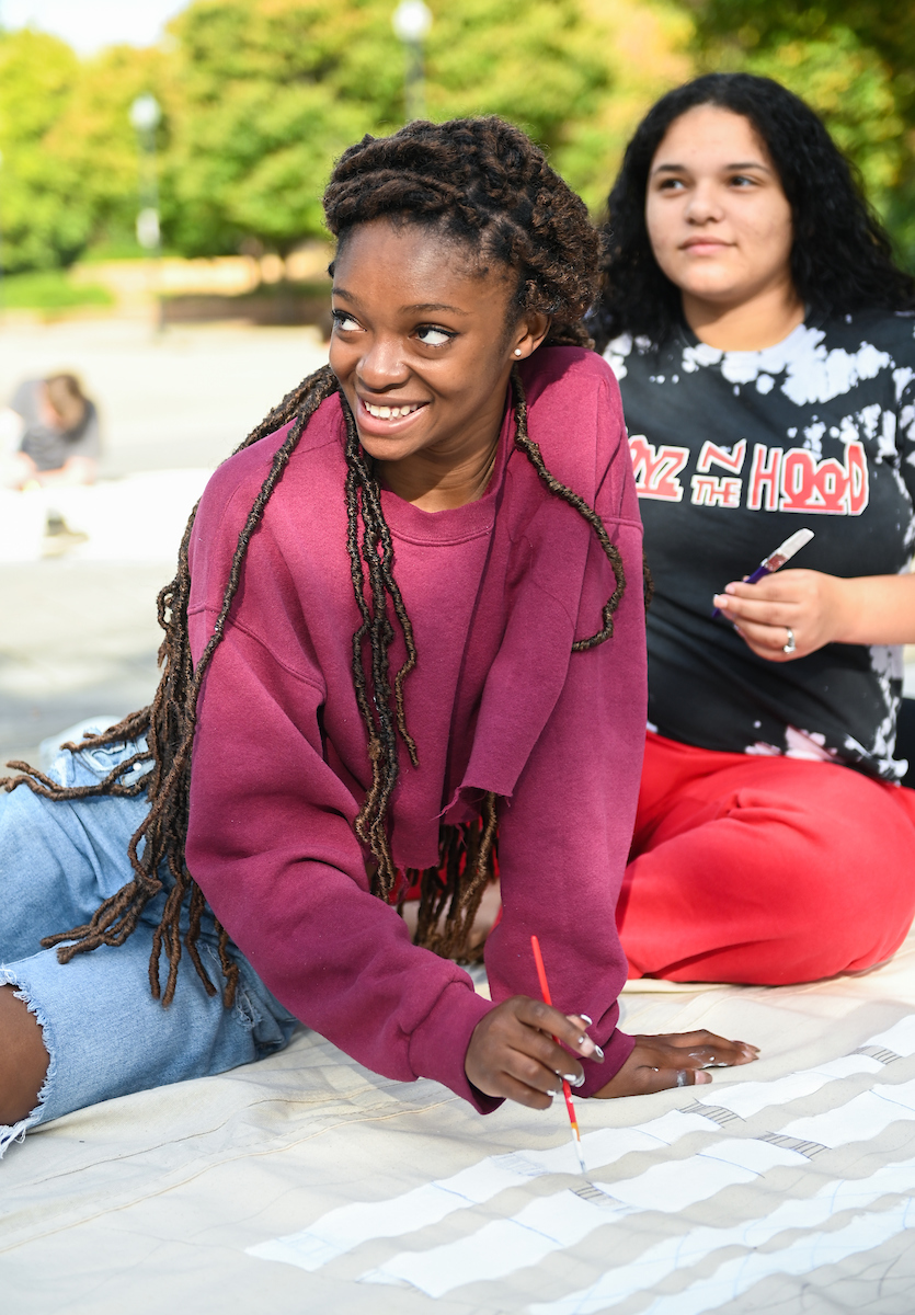 Two students sit outdoors and holds paintbrushes, one smiling while the other looks thoughtful.