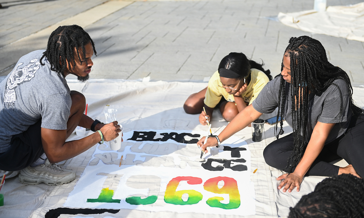 Three people are painting a large banner on the ground. The banner has colorful numbers and the word "Black".