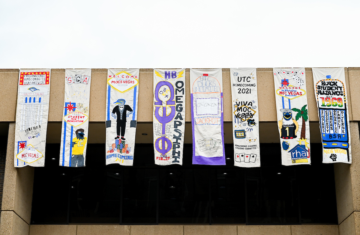 Eight colorful banners hang from a balcony, displaying creative designs for homecoming. Themes include "Mocs Vegas" and various student organizations.