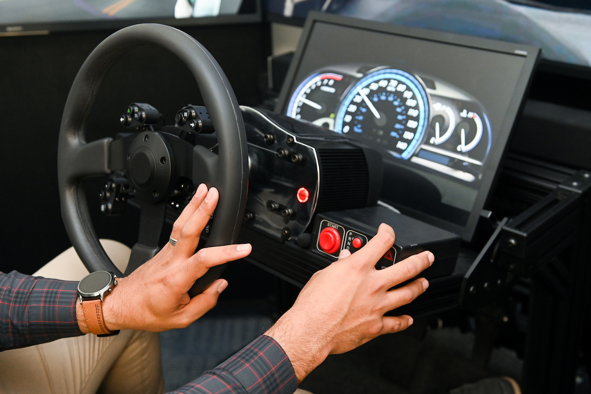 Hands on the steering wheel and control panel of a driving simulator. Dashboard shows illuminated speed and RPM gauges.
