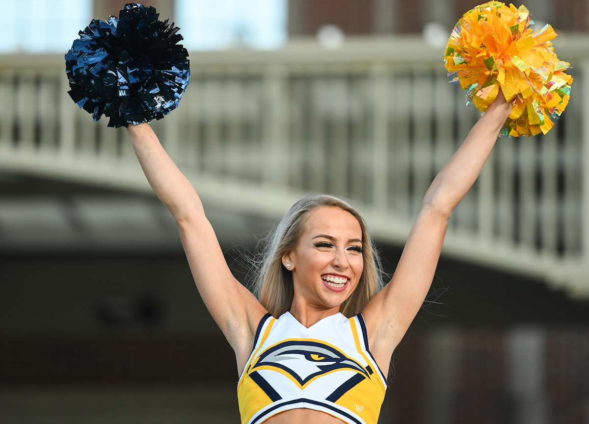 Cheerleader in yellow uniform holds blue and yellow pom-poms high, smiling energetically.