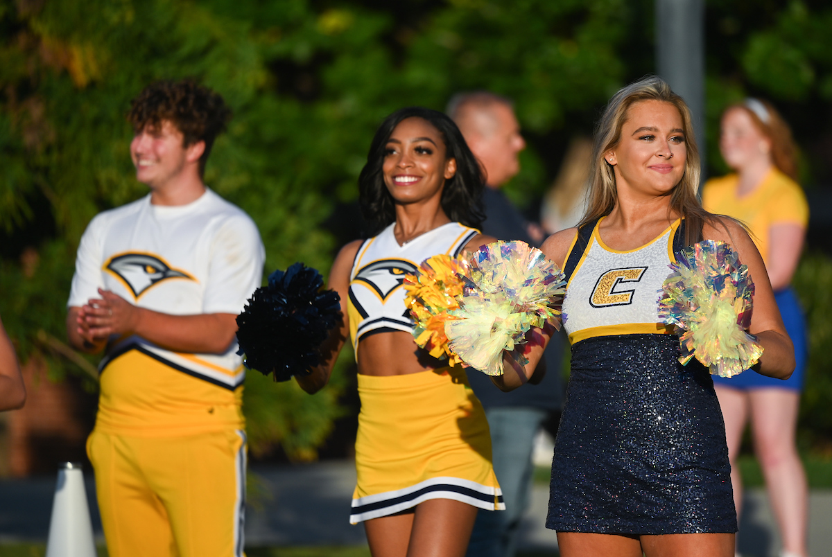 Two cheerful cheerleaders and a dancer in yellow and navy uniforms hold pom-poms. They stand outdoors, smiling confidently in the sunlight.