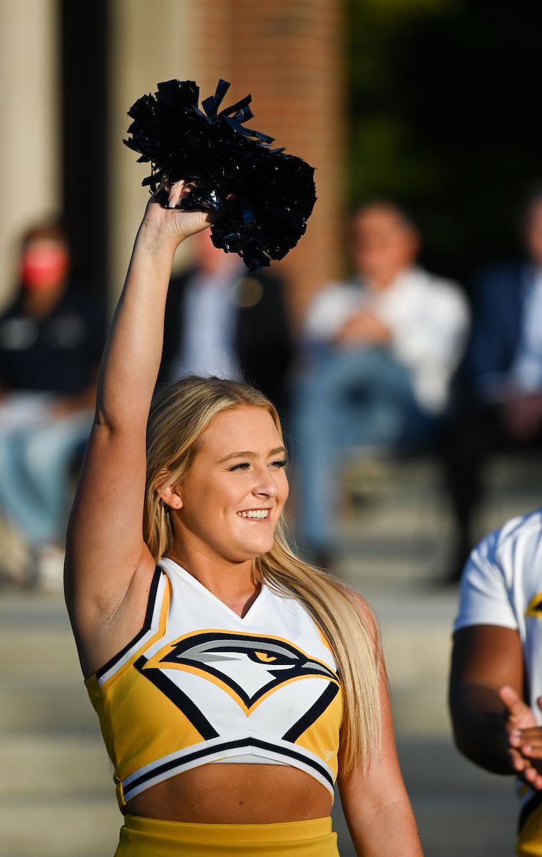 Cheerleader in yellow uniform holds a pom-pom high, smiling confidently.
