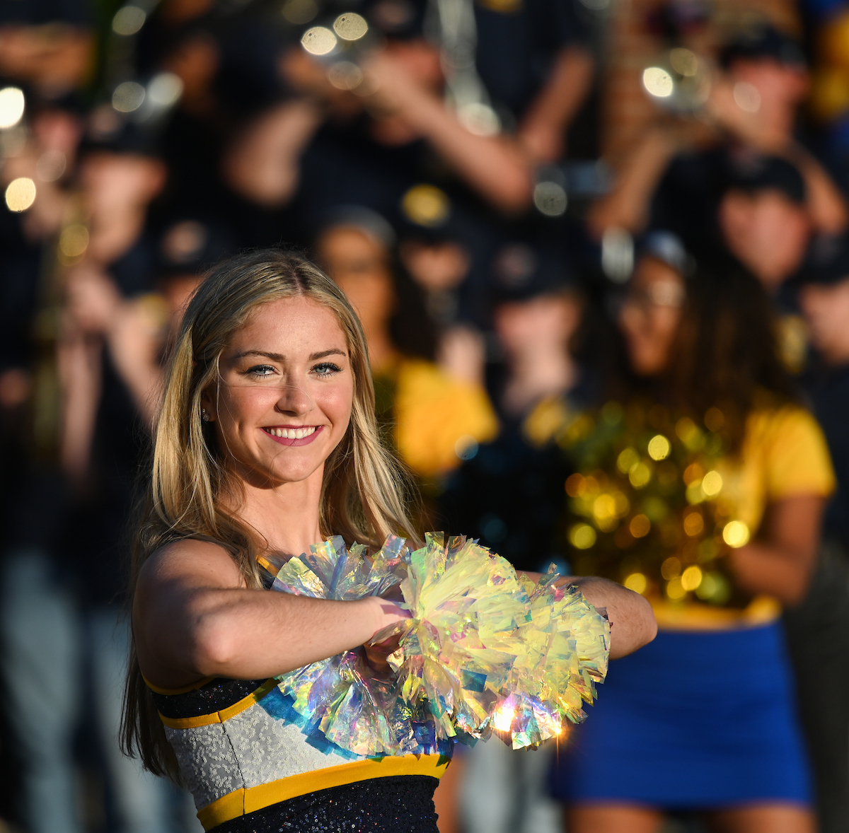 A dancer in a blue and yellow uniform smiles while holding iridescent pom-poms. A blurred crowd in similar colors is visible in the background.