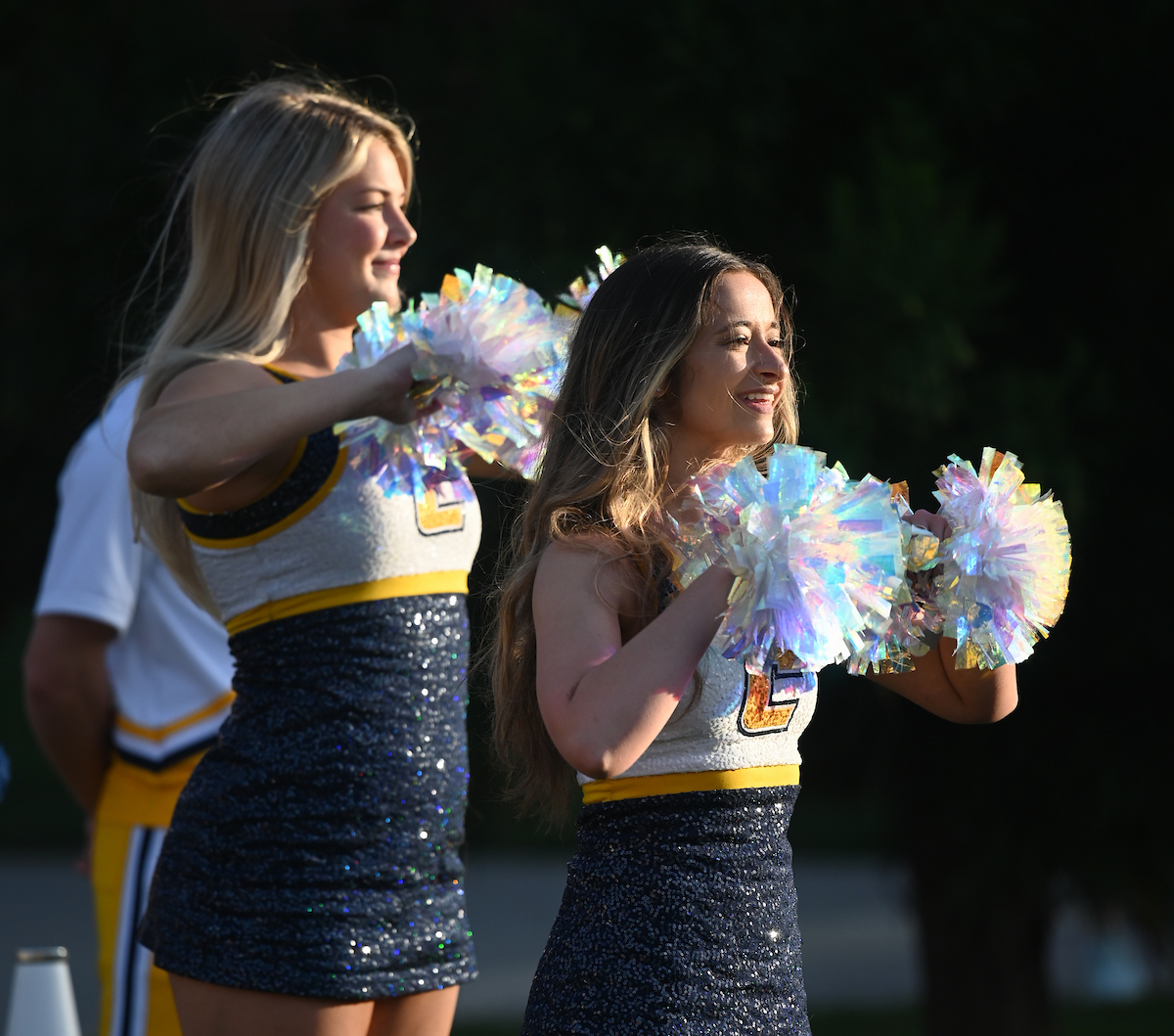 Dancers in blue and yellow uniforms hold colorful pom-poms, smiling under bright sunlight at a pep rally.