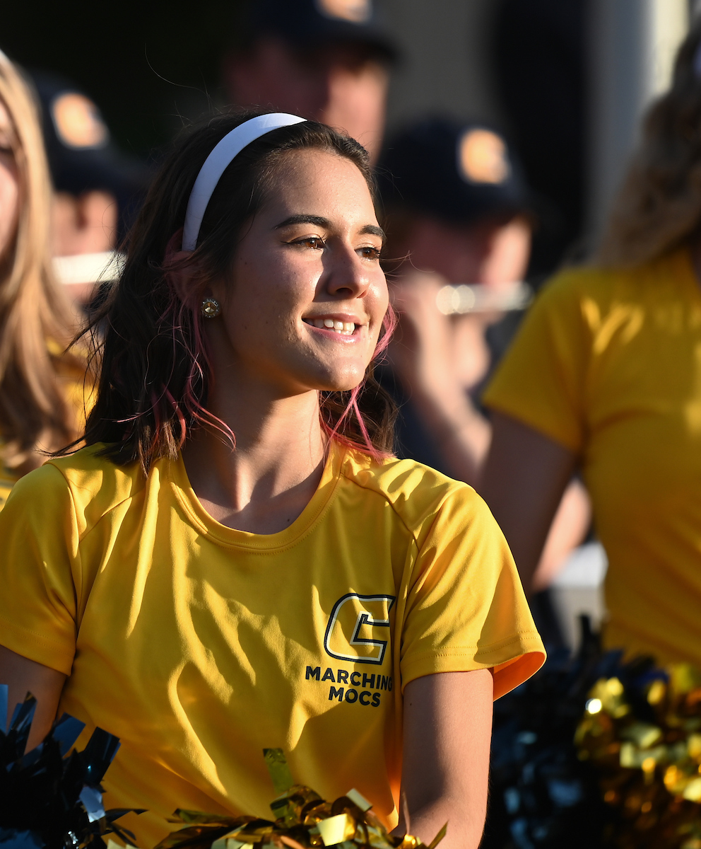 Colorguard member in a yellow "Marching Mocs" shirt smiles brightly at an outdoor event. She has a white headband and the background is slightly blurred.