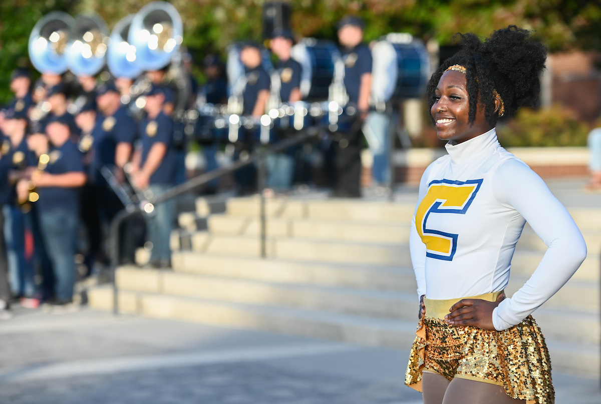 A majorette dancer in a white and gold uniform smiles confidently in the foreground, with a blurred marching band in the background.