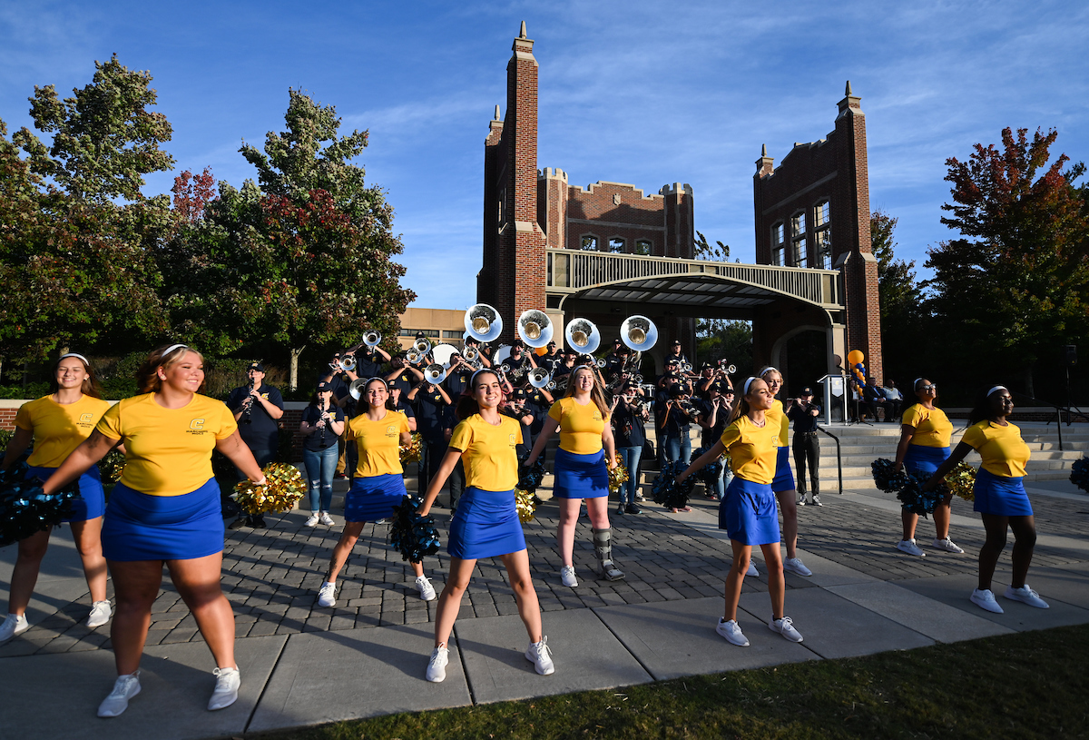 A group of colorguards in yellow tops and blue skirts perform energetically in front of a marching band with tubas, set against a historic brick building.