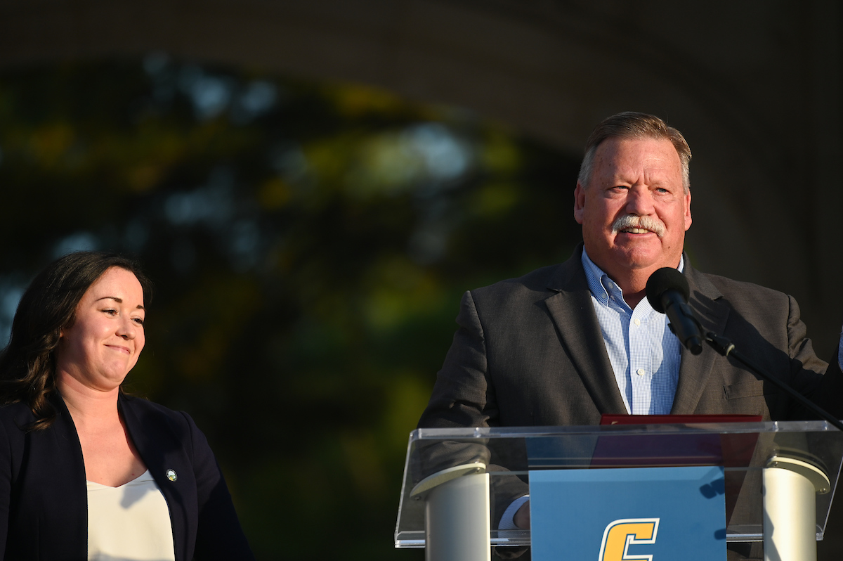 Hamilton County Mayor Jim Coppinger speaking at a podium, and Julia Bursch is standing behind him, in an outside setting.