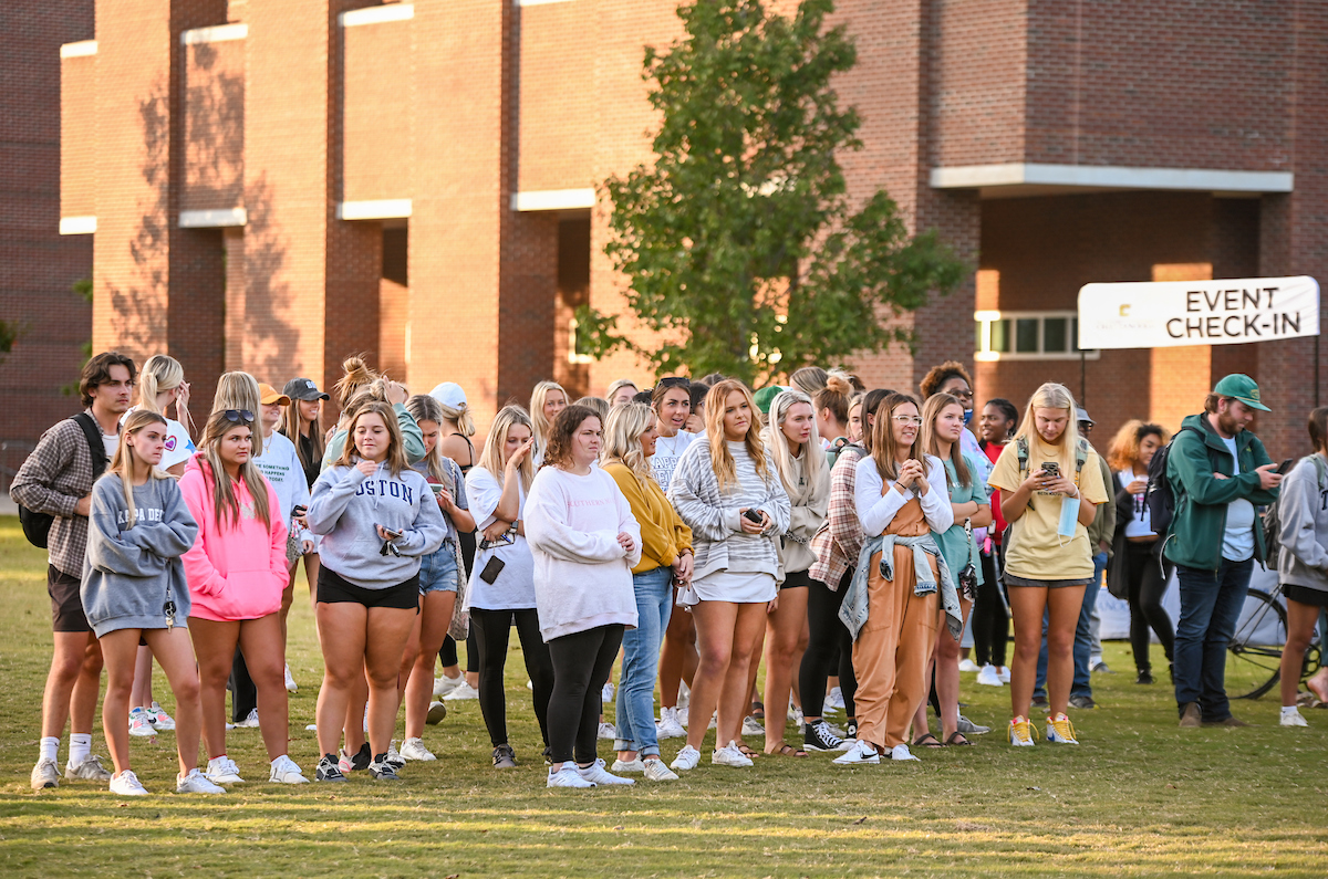 A group of students stands outside on grass in front of a brick building. A sign reads "Event Check-In."