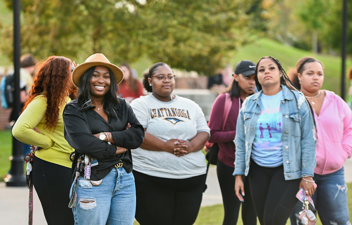 A group of students outdoors during a homecoming event, some smiling and others with neutral expressions. The background shows trees and grass.