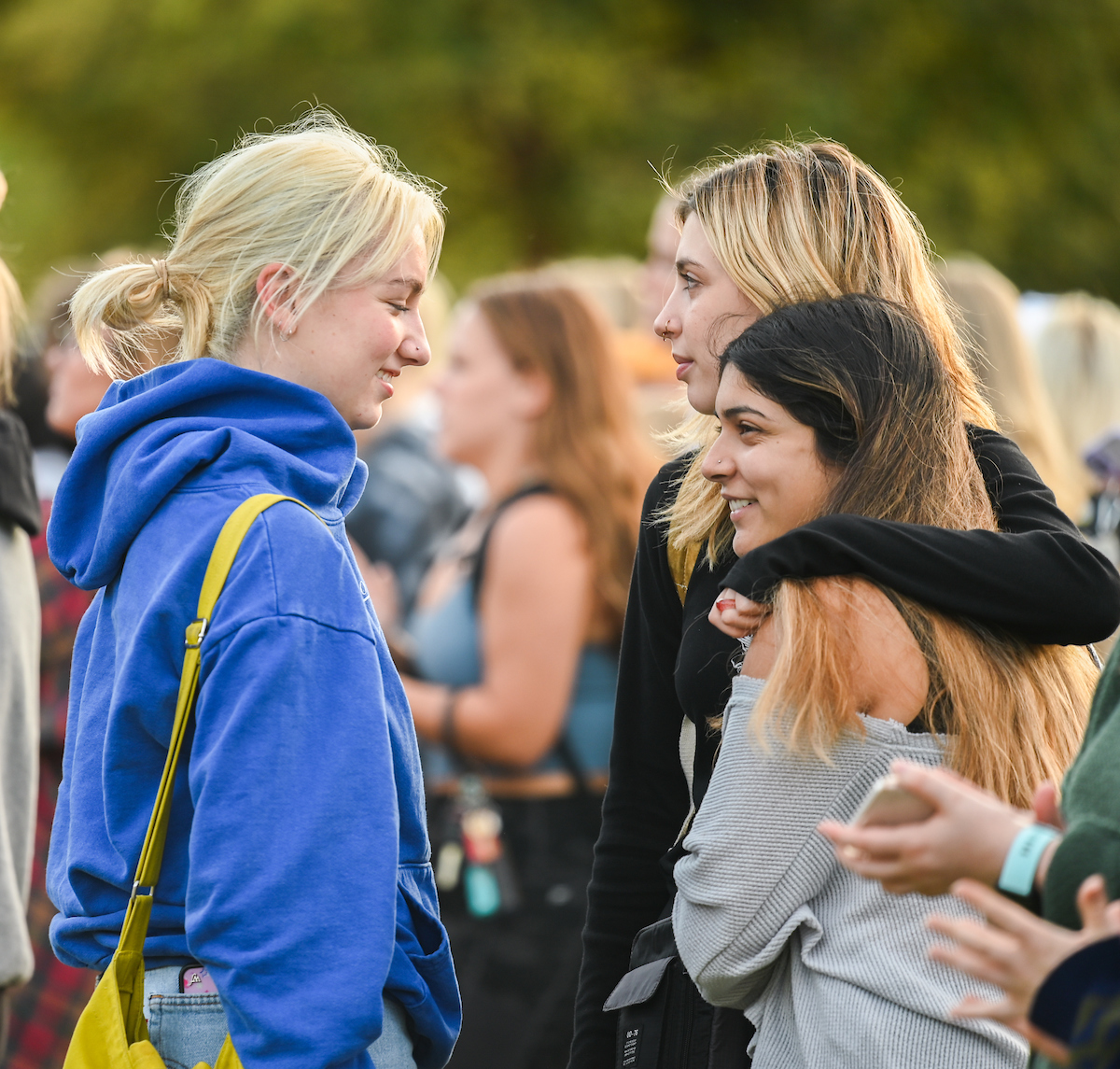 Three students stand closely together, smiling warmly in an outdoor setting.