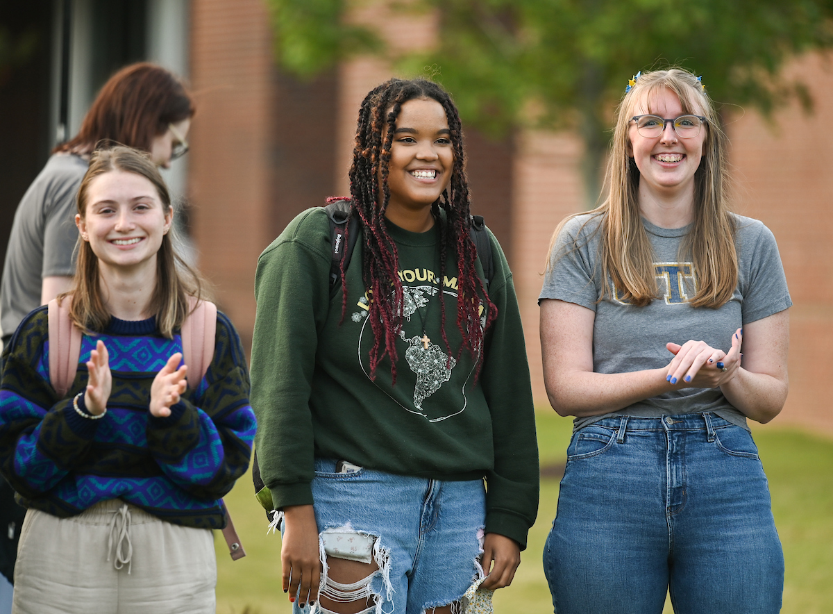 Three smiling students stand outdoors, two clap while the third has hands together.