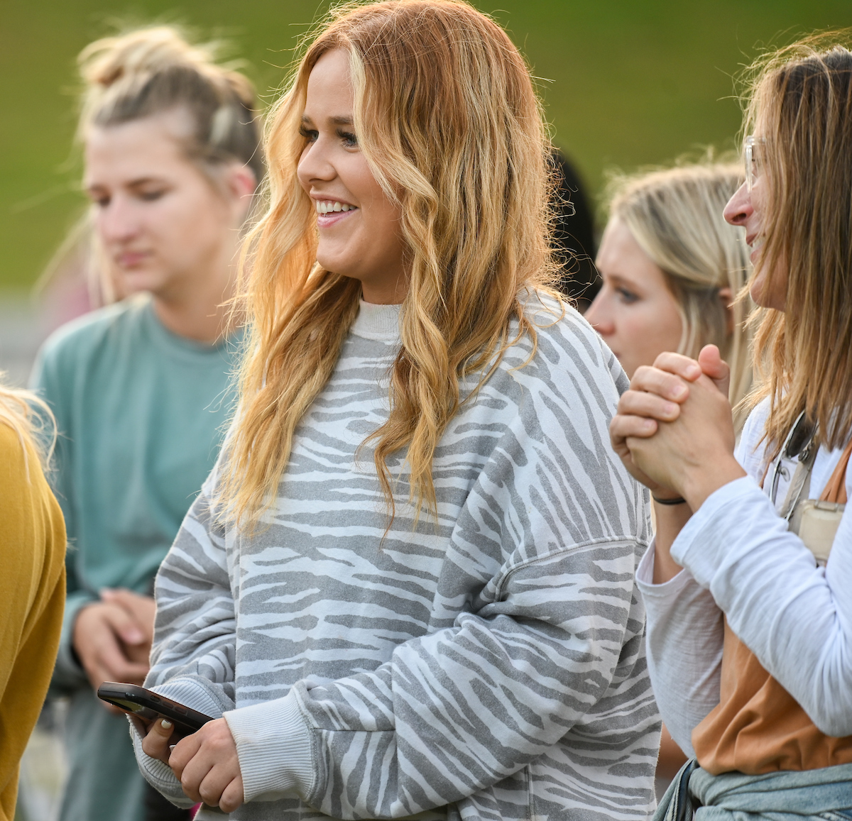 Student smiling in a gray striped sweater, holding a phone. Surrounded by people in a casual outdoor setting.