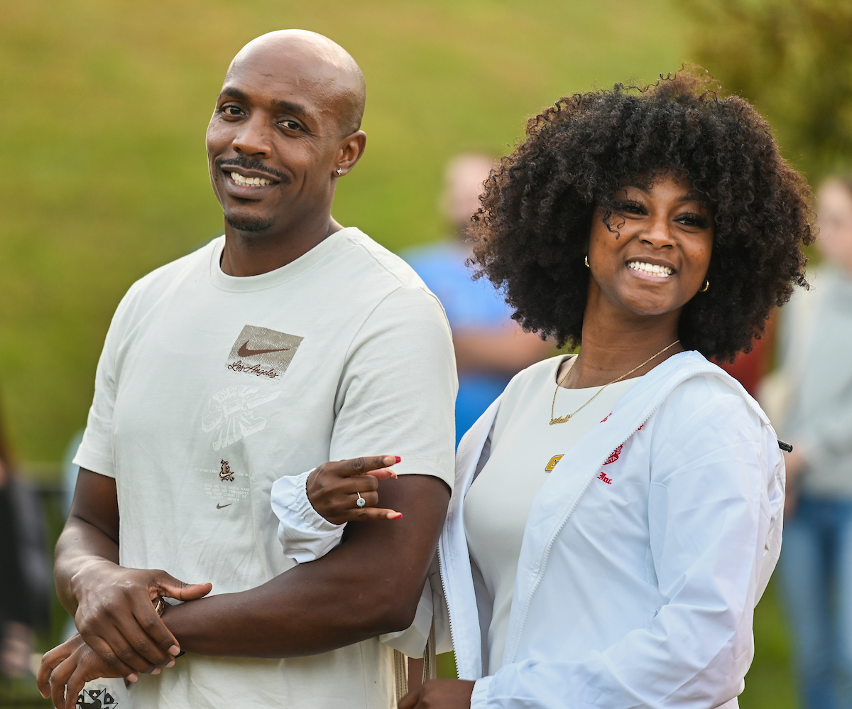 A smiling man and woman wearing casual clothes pose outdoors with others blurred in the background.