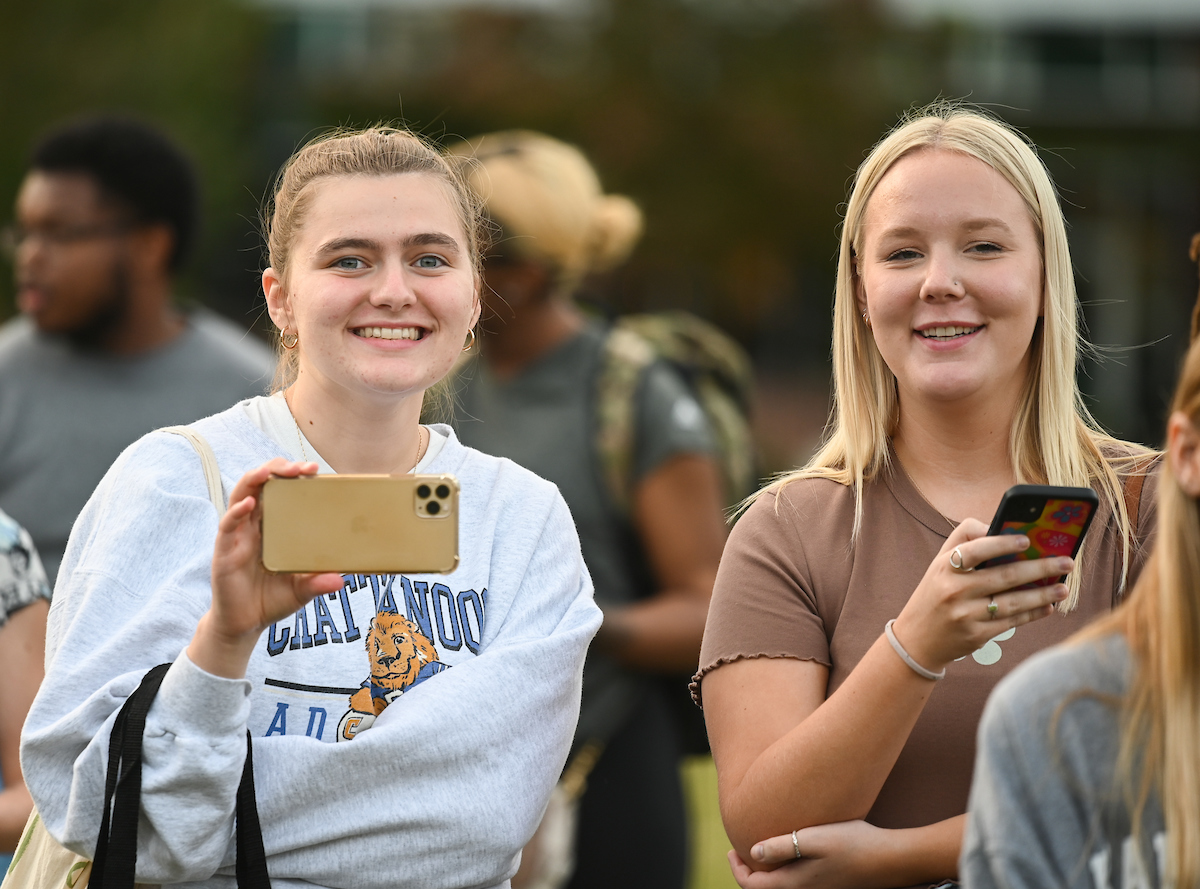 Two smiling students are taking photos with their phones at an outdoor event. They are surrounded by blurred onlookers.