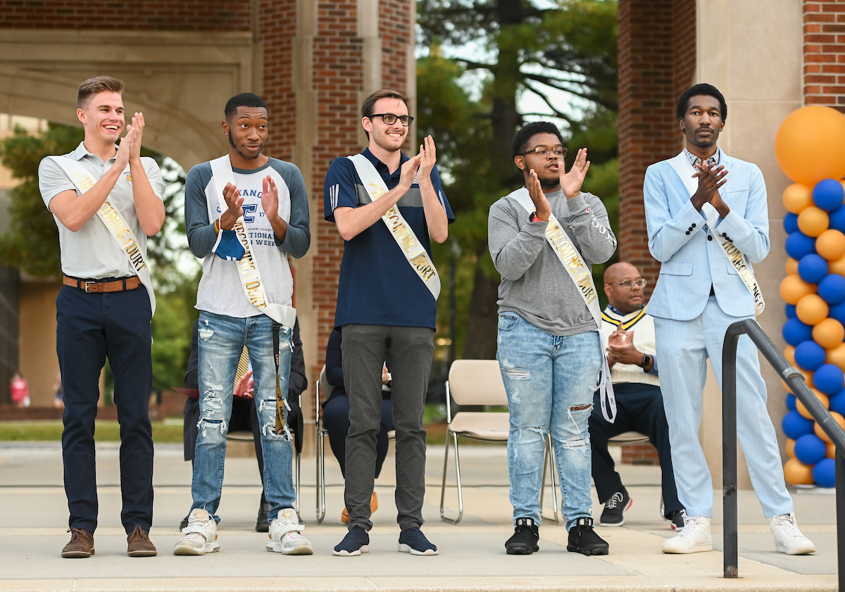 The men's Homecoming Court stands on a stage, clapping and smiling. They wear casual and formal attire, wearing homecoming sashes. The background features brick columns and blue and gold balloons.