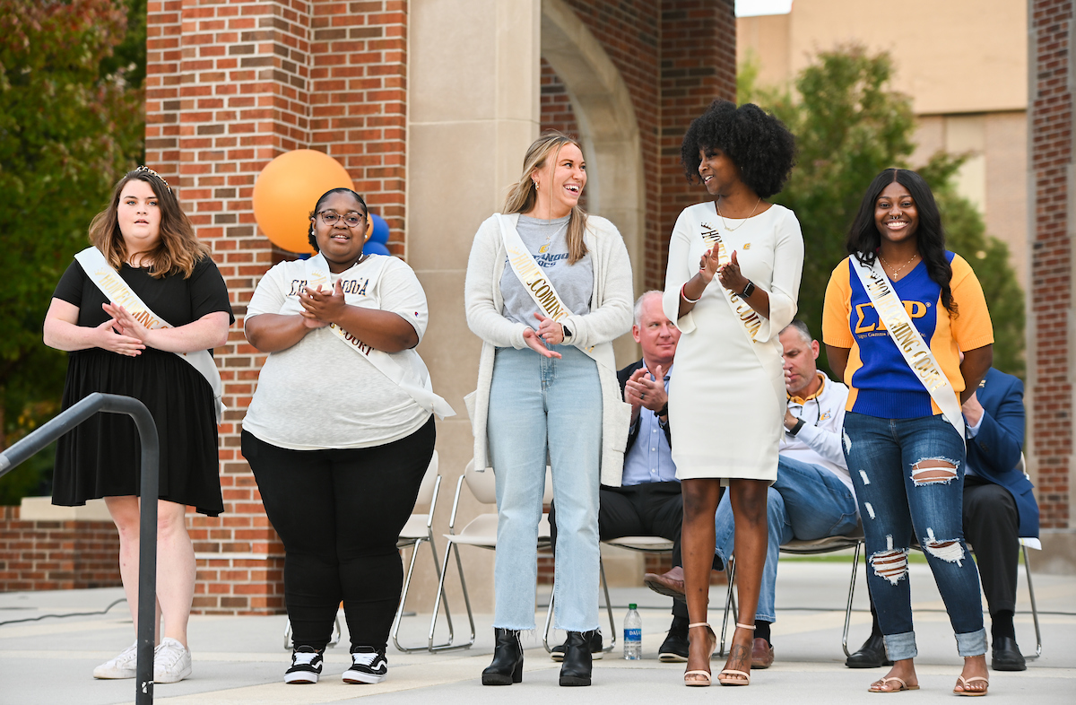 The Ladies Homecoming Court stand beside each other on an outdoor stage, smiling, some clapping. They wear "Homecoming Court" sashes.