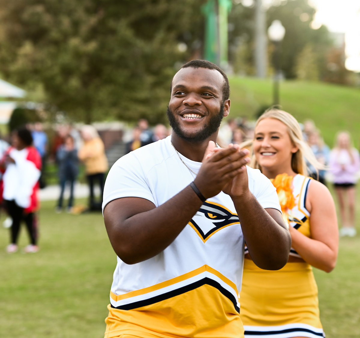 Smiling cheerleader clapping during an outdoor pep rally, wearing a white and yellow uniform.