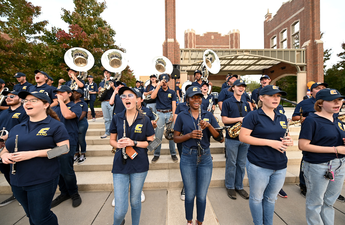 Marching Mocs play instruments on outdoor steps, wearing navy uniforms and caps.