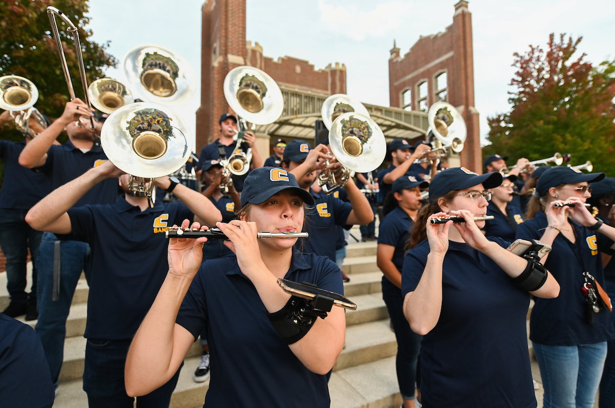 Marching Mocs play instruments on outdoor steps, wearing navy uniforms and caps.