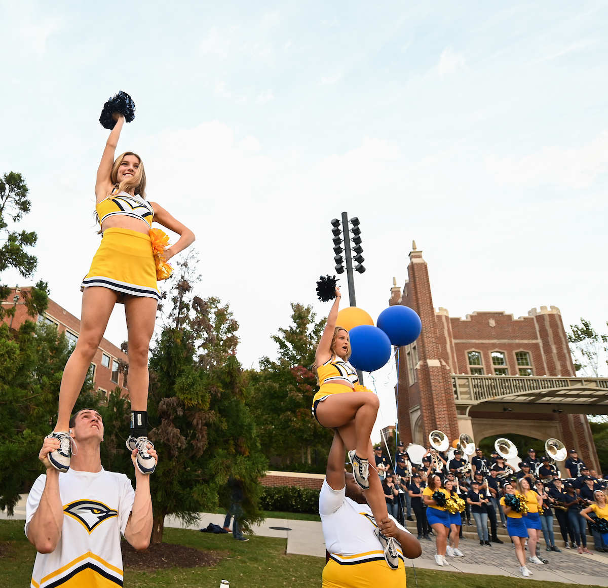 Cheerleaders in yellow and navy uniforms perform stunts with pom-poms, while a marching band is visible in the background.