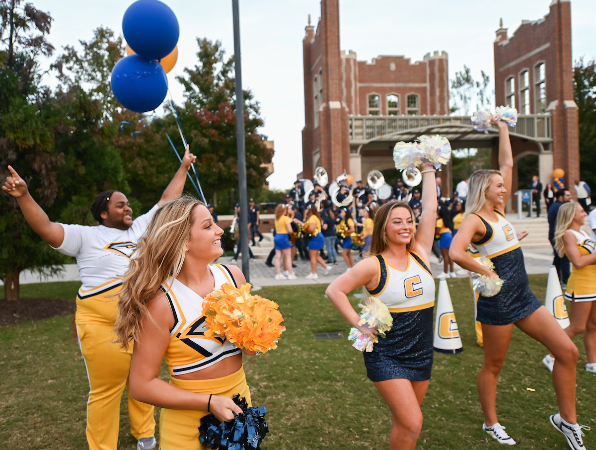 Cheerleaders and dancers in a pep rally perform energetically with pom-poms; one holds balloons. A marching band plays in the background.