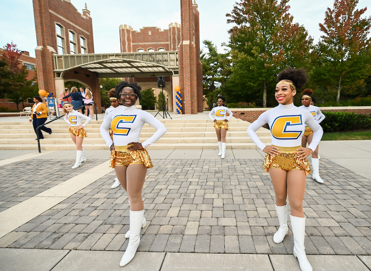 Ladies of Gold in white and gold uniforms perform outdoors in front of a red-brick building.