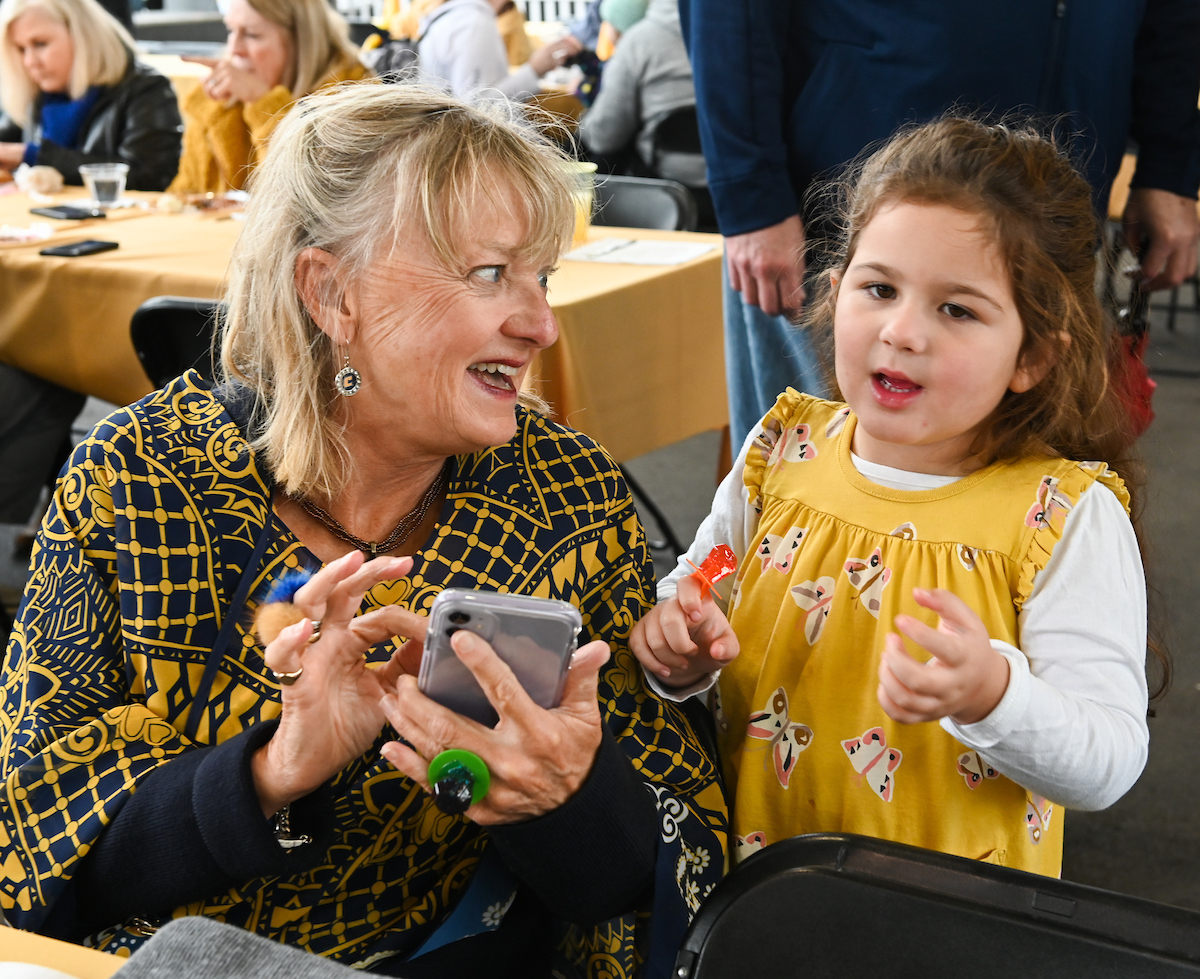 A woman and young girl smile joyfully at homecoming gathering. The woman holds a smartphone, and the girl gestures expressively.