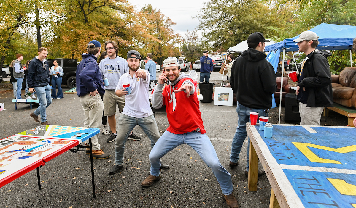 Two men in the foreground pointing and posing towards the camera. Others in the background converse at the lively outdoor gathering.