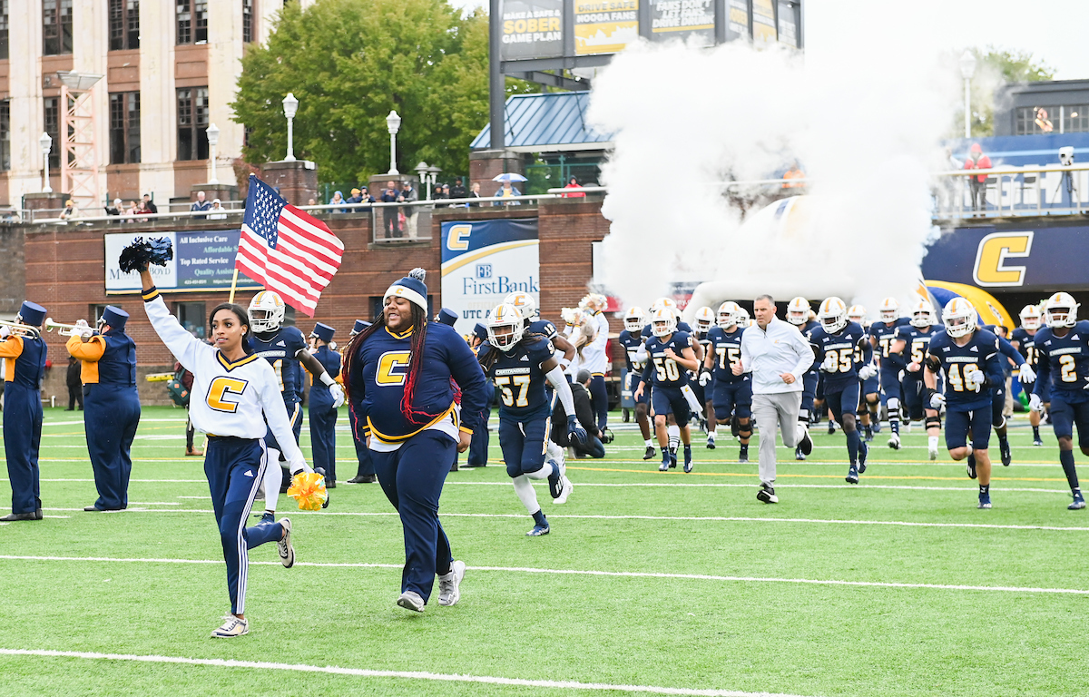 Cheerleaders and football players run energetically on the field, through smoke.