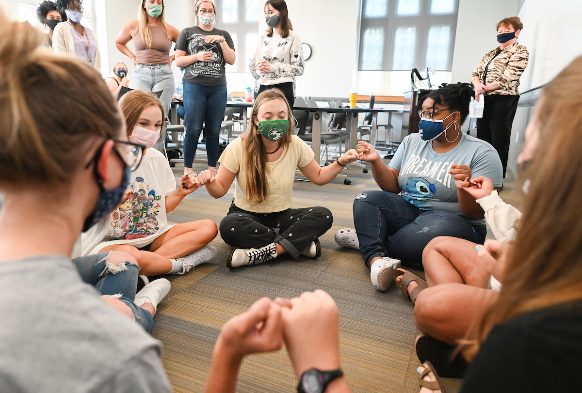 A group of students, all wearing masks, sit on a checkered floor in a circle holding pinkies.