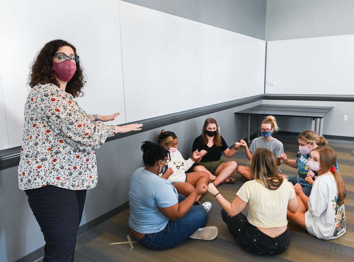 Laurie Melnik Allen, gestures toward a group of seated students, all masked, holding hands in a circle.