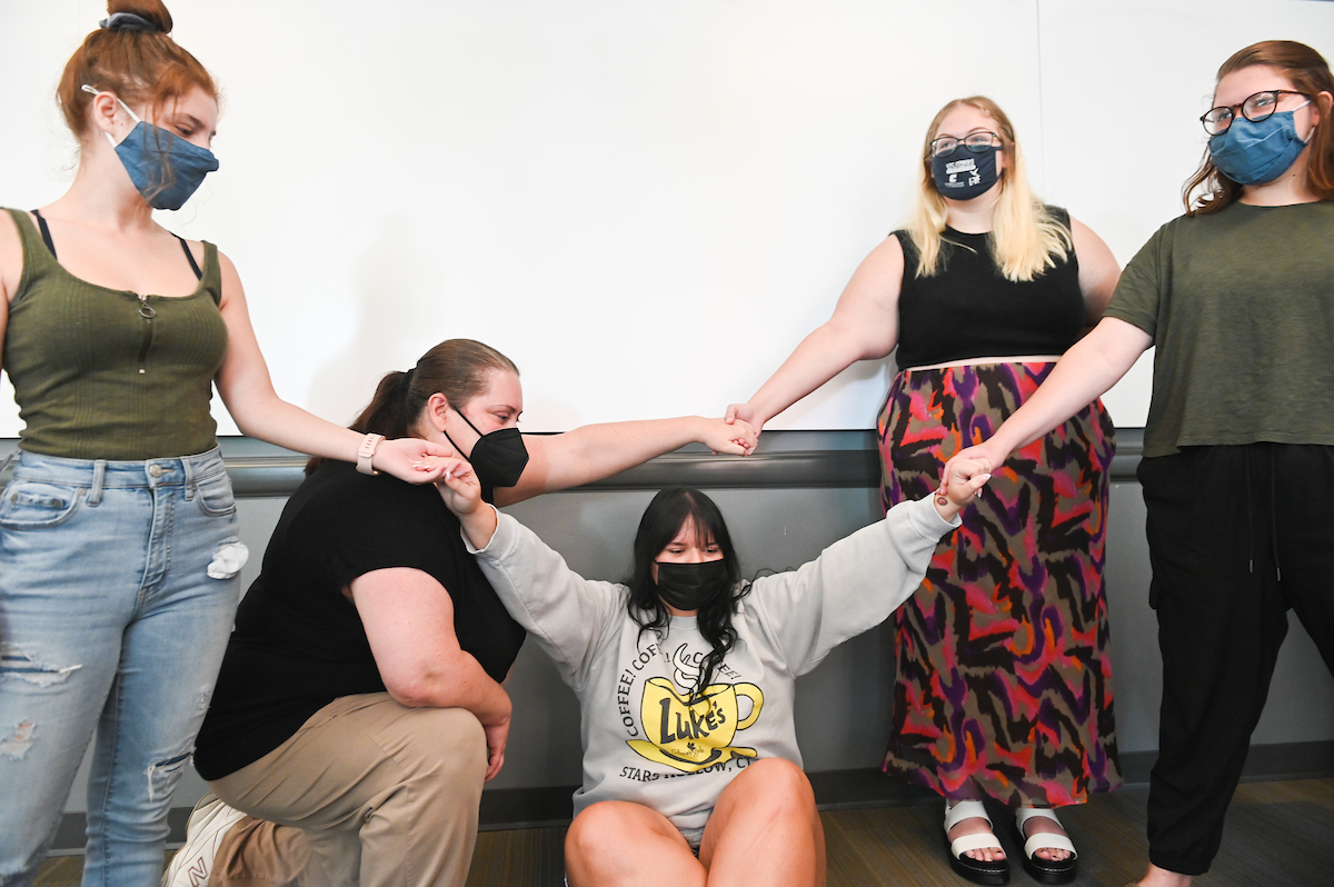 Five people, wearing masks, are in a classroom. One person is kneeling with arms raised, while the others stand in a circle, holding hands.