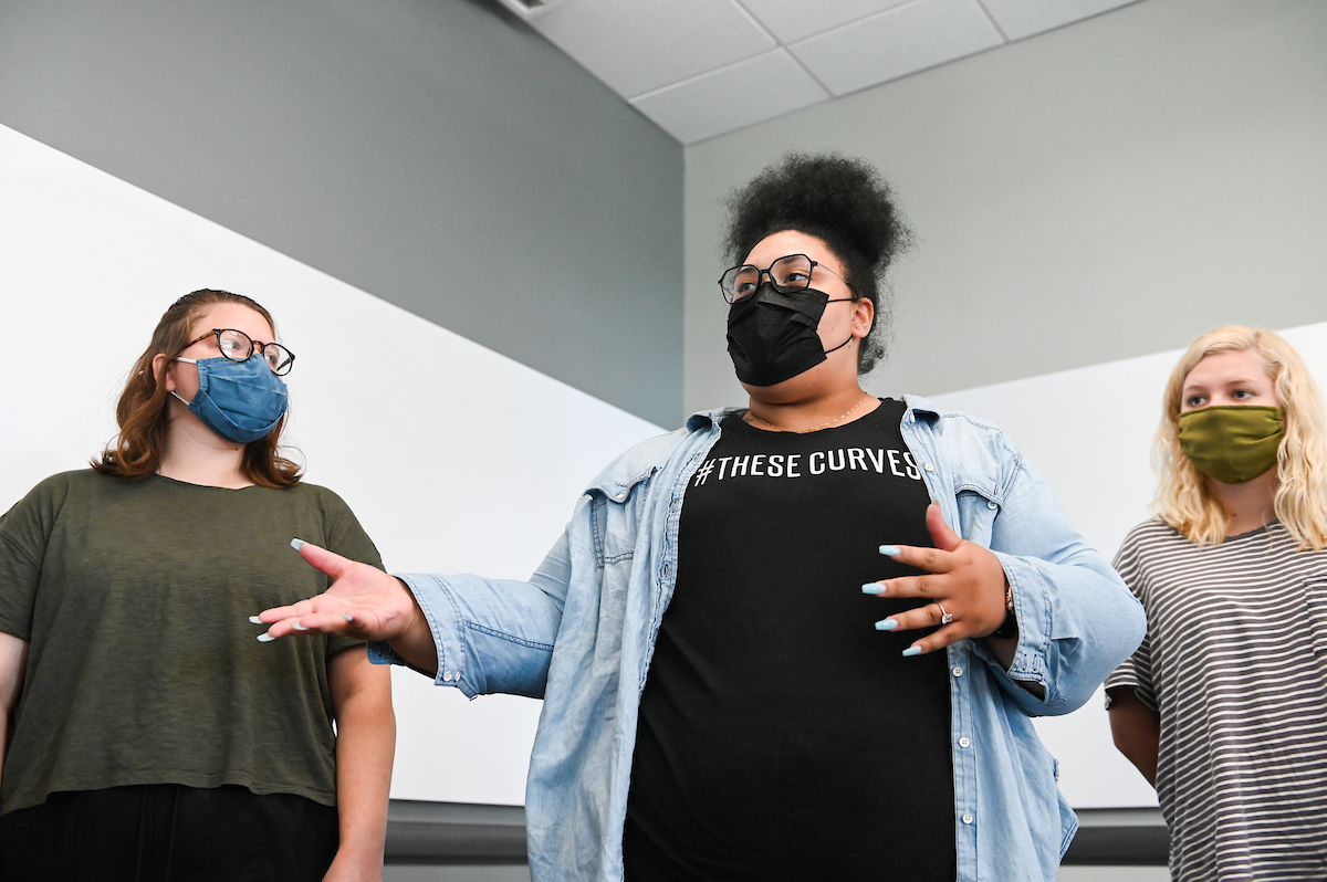 Three women wearing face masks stand in a room with a gray and white wall.